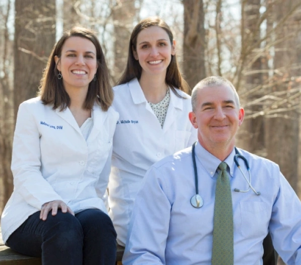 Group portrait of three veterinary staff members, two women in white coats and a man in a shirt and tie with a stethoscope, smiling outdoors